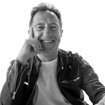 Gray scale portrait of a smiling man with short hair touching his face while wearing a dark textured jacket
