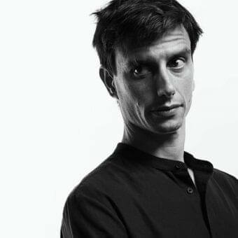 Monochromatic portrait of a man facing forward with short dark hair and a dark collarless garment against a white background
