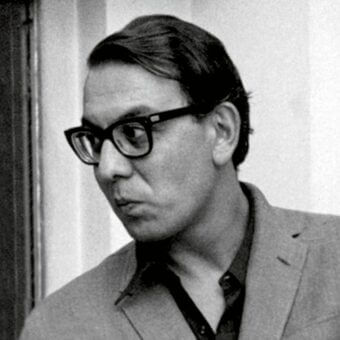 Portrait of Giampiero Vitelli wearing black eyeglasses and a light blazer, facing left with a neutral expression against a pale wall.