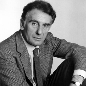 Man with short dark hair wearing a checkered jacket, white shirt, and striped tie looks forward with a serious expression