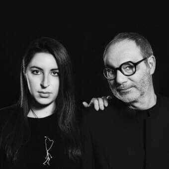 Grayscale portrait of a woman with long dark hair and a man with short gray hair, both facing forward with direct gazes against a dark backdrop