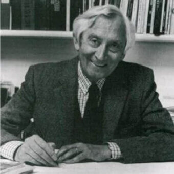 Seated person with light hair smiles toward the camera behind a flat surface with hands resting on papers before a filled bookshelf