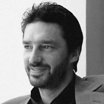 Grayscale portrait of a man with dark hair and beard looking upwards while wearing a dark collared shirt and light jacket