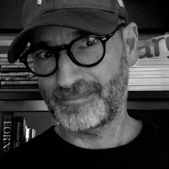 Monochrome portrait of a man with facial hair and glasses, facing forward with stacked rectangular objects and blurred lettering behind him