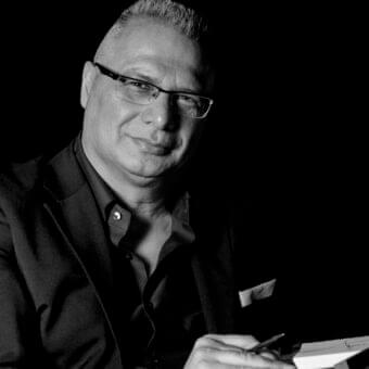 Grayscale portrait shows a man with glasses holding a pen over white paper against a black background