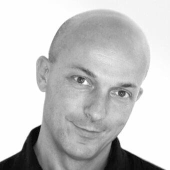 Grayscale portrait of a hairless man facing slightly right with a subtle smile and dark clothing against a light background