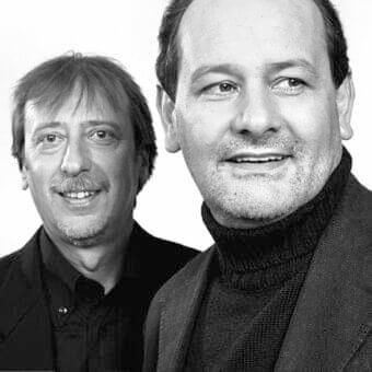 Grayscale portrait of two men facing forward with stubble and styled hair wearing dark collared garments against a light background