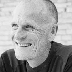 Smiling man with graying hair and visible facial lines against a blurred gray brick background