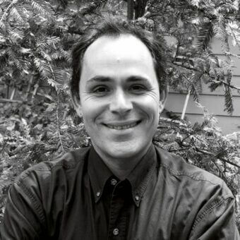 Portrait of James Rosen, smiling, wearing a dark brown shirt, against blurred evergreen foliage and building backdrop in natural daylight.