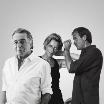 Three people are shown in close-up, a seated man, a standing woman with glasses, and another man adjusting her hair