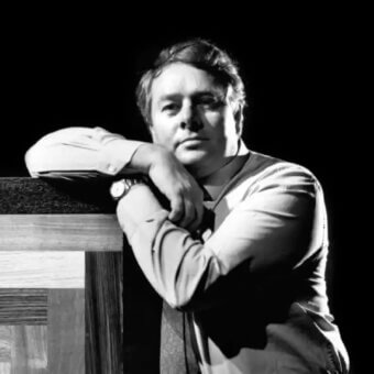 Man leans on stacked rectangular planes with visible texture, wearing a light shirt, dark tie and wristwatch against a dark background