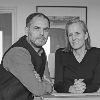 Black and white portrait of two people seated at a white table, with framed artwork reading "FOREST TOWNHOUSE FORREN MECHLER" in the background.