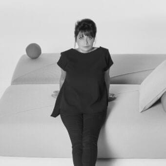 Person with updo stands before a grayscale curved structure with a cushion and spherical object above