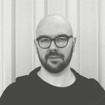 Bald man with dark beard and round glasses looks forward against a light ribbed vertical backdrop