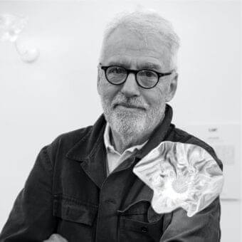 Older man with white beard and glasses holds a crumpled metallic object against a light wall