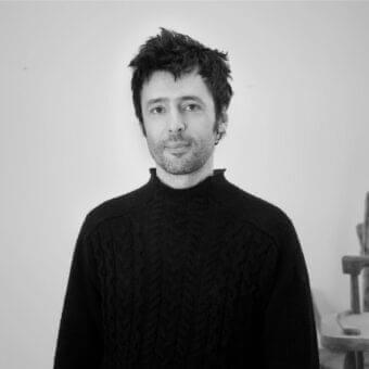 Monochromatic portrait of a man with dark hair wearing a dark knit garment against a light gray background with stacked dark shapes