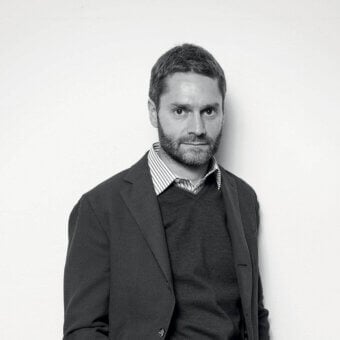Grayscale portrait of a bearded man wearing a dark jacket, sweater and striped shirt, looking directly forward against a plain backdrop