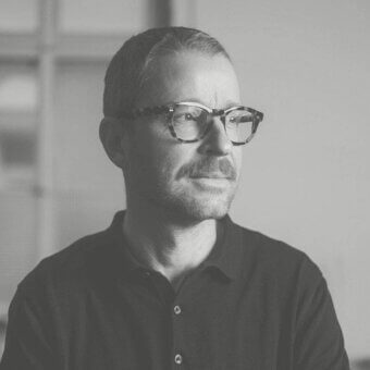 Grayscale portrait of a man with short dark hair, glasses, mustache and beard looking to the right against a blurred architectural background