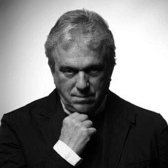 Monochromatic portrait of a man with gray hair in a dark jacket looking down with a hand raised to his chin