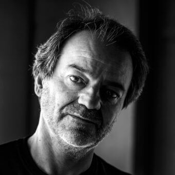 Black and white portrait of Marco Zito, a man with graying hair and beard, wearing a dark t-shirt, against a blurred dark background.