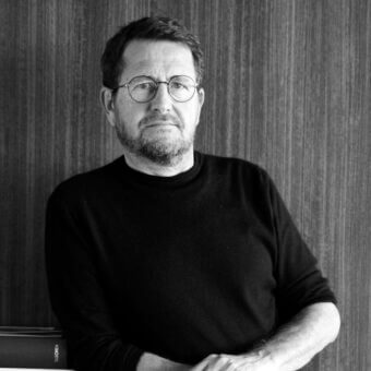 Black and white portrait of Mario Mazzer, a man with glasses, crossed arms, and a book titled “Sachsen”, leaning against a textured wall.