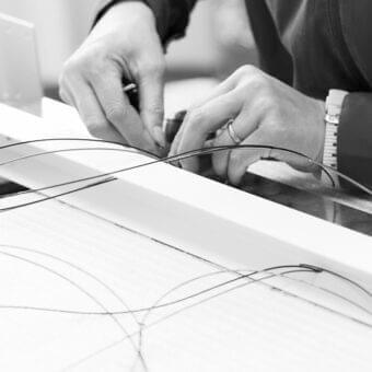 Hands manipulate dark flexible lines on a large white surface with a pen and metallic wristwatch visible
