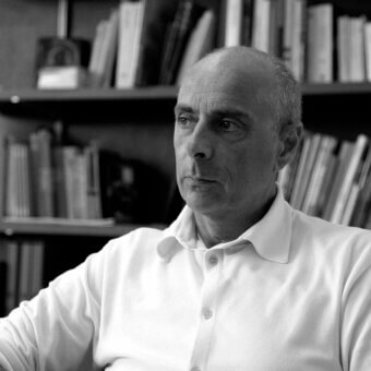 Grayscale portrait of a man with a shaved head looking right, bookshelves filling the background and a dark rectangle visible to his left