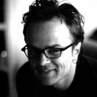 Monochrome portrait of a person with short dark hair, glasses, and a slight smile, facing left against a blurred background