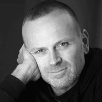 Man with shaved head and stubble rests chin on hand, face turned toward viewer in grayscale lighting