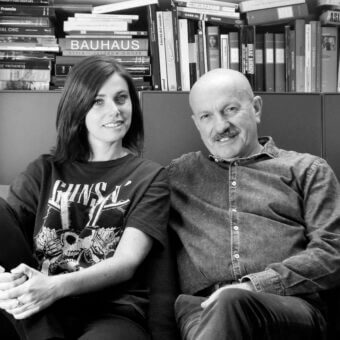 Two people seated on maroon armchair, bookshelves backdrop, woman in graphic tee, man in plaid shirt, soft diffused lighting.