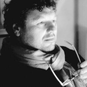 Grayscale portrait of a man in profile with curly hair, stubble, and glasses held in hand looking right