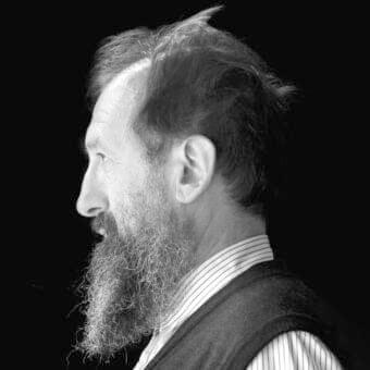 Monochrome portrait of a man with a beard facing left, wearing a striped collared shirt and dark vest against a dark background