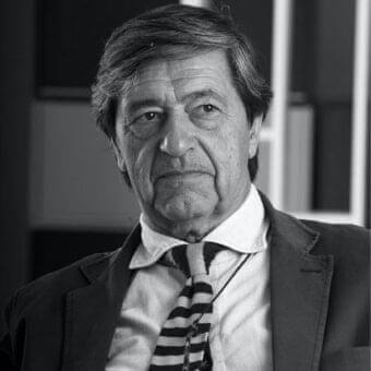 Gray scale portrait of a man in a suit jacket, white shirt and striped tie wearing suspended eyeglasses with blurred architectural background