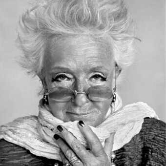 Close-up portrait of a person with spiky hair, glasses, rings, and a draped fabric around the neck