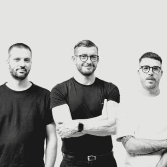 Monochromatic portrait of three men with short hair and facial hair wearing dark t-shirts against a pale gray backdrop