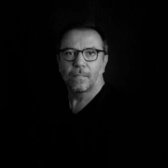 Grayscale portrait of a person with short gray hair and glasses, wearing a dark v-neck top against a black background