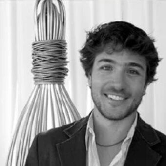 Grayscale portrait of a smiling man with curly hair beside a tall conical structure of parallel metallic elements against a pale backdrop