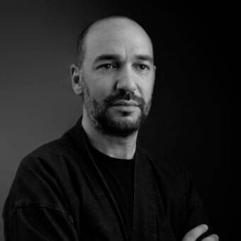 Grayscale portrait of a man with a shaved head and beard, arms crossed, wearing a dark collared shirt against a gradient background
