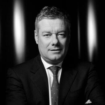 Middle-aged man in a dark pinstriped suit and patterned tie looks forward with blurred vertical shapes behind him