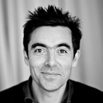 Centered black and white portrait shows a smiling man with textured dark hair and visible facial lines against a pale striated background