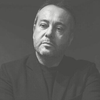 Grayscale portrait of a man facing left with short hair, beard, and a textured dark jacket against a plain gray background