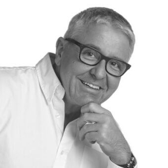 Pierangelo Sciuto portrait, black and white, man with gray hair, glasses, white shirt, hand to chin, direct gaze, soft lighting.