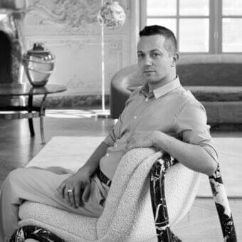 Seated person with ring wears light clothing near a chair with textured seat and dark patterned frame in a room with pale flooring
