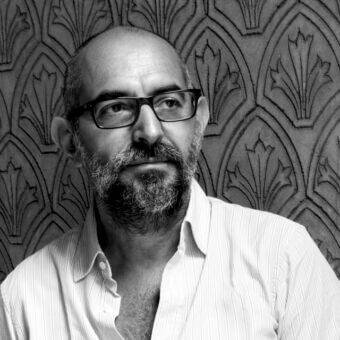 Black and white portrait of Pietro Ferruccio Laviani, a man with beard and glasses, wearing a striped shirt, against a patterned wall.