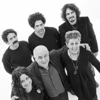 Six people stand and sit closely together against a neutral gray backdrop with overlapping arms
