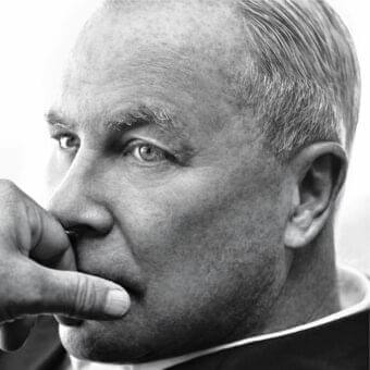 Middle-aged man’s graying profile faces slightly left with a finger near his mouth against a light gray backdrop