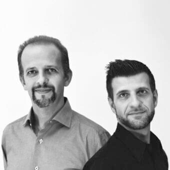 Monochromatic portrait of two men facing forward with beards, wearing collared shirts against a light gray backdrop