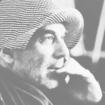 Monochrome portrait of a man in profile wearing a patterned cap, hand raised touching chin, with blurred dark background tones