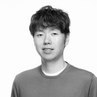Black and white portrait of a man with layered dark hair wearing a gray sweatshirt and white t-shirt, neutral expression, plain background.