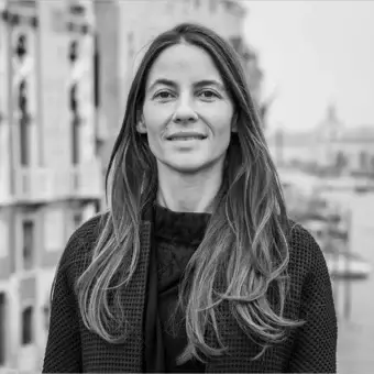 Grayscale portrait of a woman with wavy hair wearing a dark jacket before a blurred cityscape with columns and distant boats